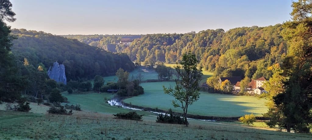 Ein malerisches Tal mit einem sich schlängelnden Fluss, grünen Feldern, verstreuten Bäumen, bewaldeten Hügeln und ein paar Häusern, die in der Morgensonne unter einem klaren blauen Himmel liegen.