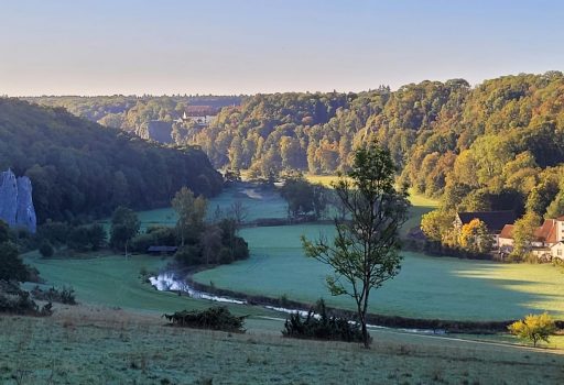 Ein malerisches Tal mit einem sich schlängelnden Fluss, grünen Feldern, verstreuten Bäumen, bewaldeten Hügeln und ein paar Häusern, die in der Morgensonne unter einem klaren blauen Himmel liegen.