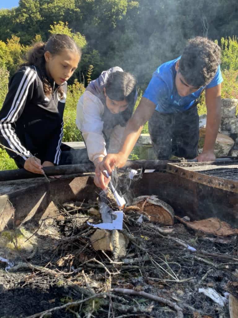 Drei Kinder beugen sich an einem sonnigen Tag über eine Feuerstelle im Freien und benutzen Stöcke, um Papier und Zweige zu räuchern, mit Bäumen und Grün im Hintergrund.