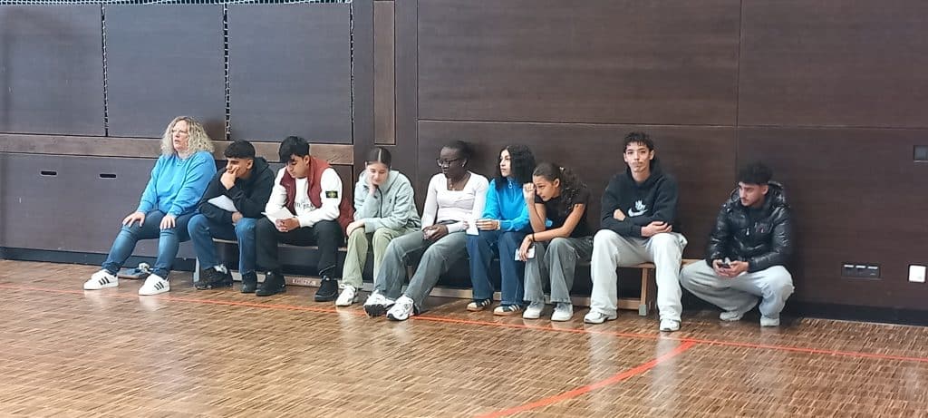 Neun Personen, darunter Männer und Frauen, sitzen auf einer Bank vor einer dunklen Holzwand in einer Turnhalle. Die meisten sehen entspannt oder konzentriert aus, einige benutzen ihre Telefone, und eine Person trägt eine leuchtend blaue Jacke.