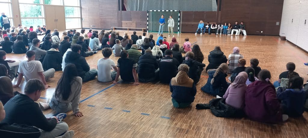 Eine große Gruppe von Schülern sitzt auf dem Boden einer Turnhalle, gegenüber stehen und sitzen einige Personen auf Bänken in der Nähe eines Torpfostens, im Hintergrund sind Holzwände und große Fenster zu sehen.
