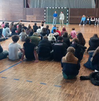 Eine große Gruppe von Schülern sitzt auf dem Boden einer Turnhalle, gegenüber stehen und sitzen einige Personen auf Bänken in der Nähe eines Torpfostens, im Hintergrund sind Holzwände und große Fenster zu sehen.