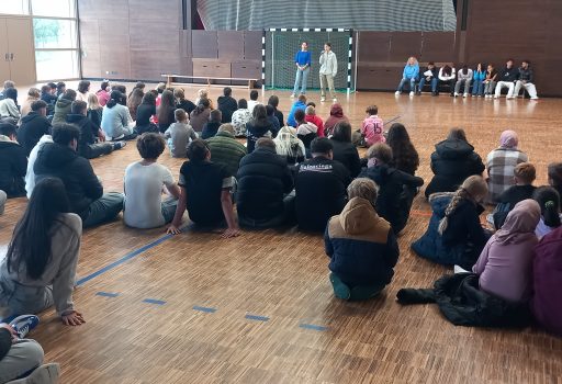 Eine große Gruppe von Schülern sitzt auf dem Boden einer Turnhalle, gegenüber stehen und sitzen einige Personen auf Bänken in der Nähe eines Torpfostens, im Hintergrund sind Holzwände und große Fenster zu sehen.