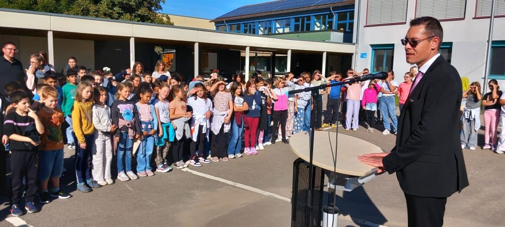 Ein Mann im Anzug spricht auf einem Podium im Freien vor einer großen Gruppe von Kindern und Erwachsenen, die in Reihen auf einem Schulhof stehen. Das Wetter ist sonnig und das Schulgebäude ist im Hintergrund zu sehen.