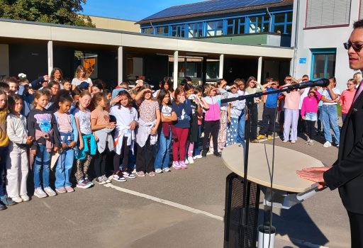 Ein Mann im Anzug spricht auf einem Podium im Freien vor einer großen Gruppe von Kindern und Erwachsenen, die in Reihen auf einem Schulhof stehen. Das Wetter ist sonnig und das Schulgebäude ist im Hintergrund zu sehen.