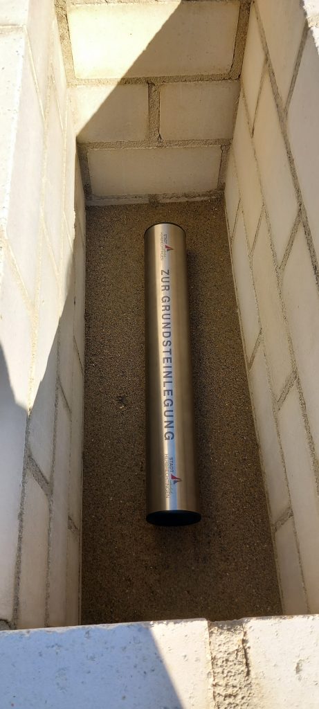 Eine metallene Zeitkapsel mit der Aufschrift ZOB GRUNDSTEINLEGUNG liegt auf Sand in einer rechteckigen, mit weißen Ziegeln ausgekleideten Grube.