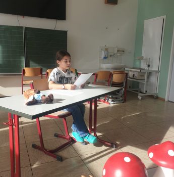 Ein Kind sitzt allein an einem Schreibtisch in einem hellen Klassenzimmer und liest von einem Blatt Papier ab. Auf einem Tisch in der Nähe befinden sich Holzdekorationen, darunter rote Pilze und ein Igel. Im Hintergrund sind leere Stühle und eine Tafel zu sehen.