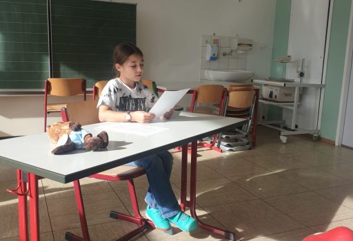 Ein Kind sitzt allein an einem Schreibtisch in einem hellen Klassenzimmer und liest von einem Blatt Papier ab. Auf einem Tisch in der Nähe befinden sich Holzdekorationen, darunter rote Pilze und ein Igel. Im Hintergrund sind leere Stühle und eine Tafel zu sehen.