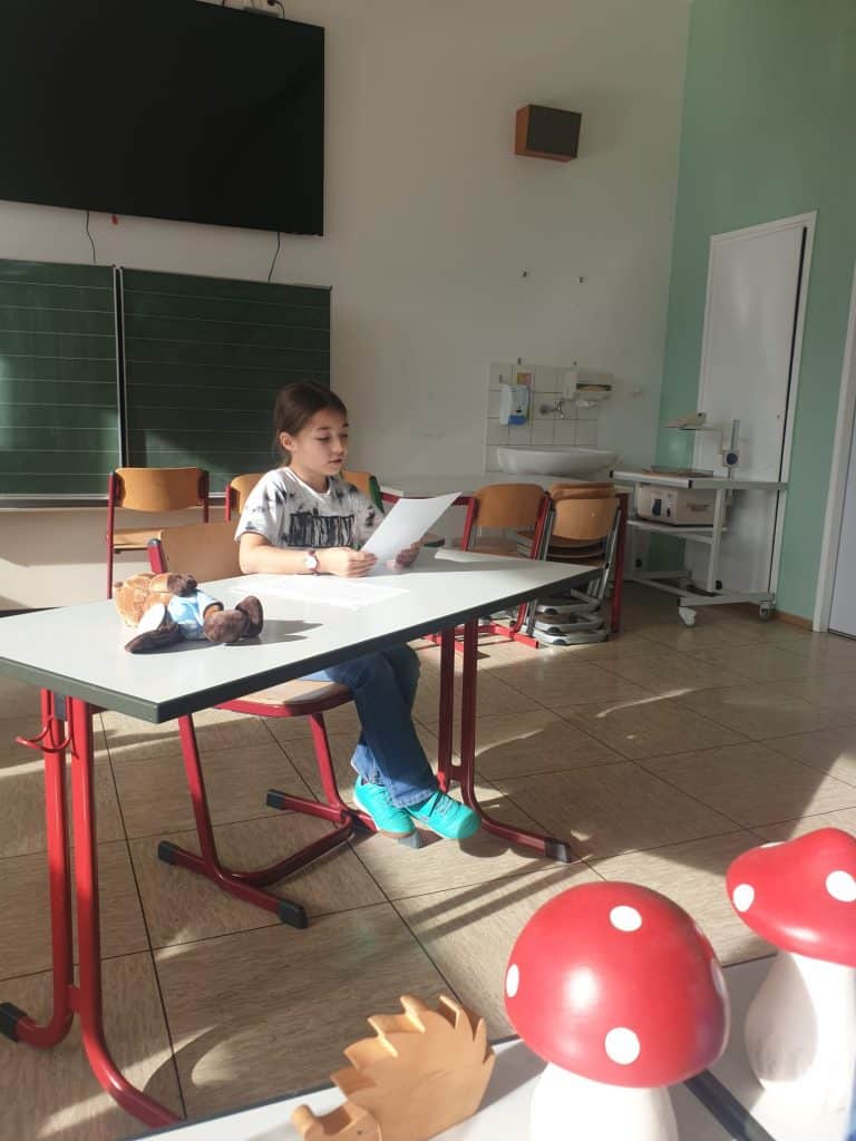 Ein Kind sitzt allein an einem Schreibtisch in einem hellen Klassenzimmer und liest von einem Blatt Papier ab. Auf einem Tisch in der Nähe befinden sich Holzdekorationen, darunter rote Pilze und ein Igel. Im Hintergrund sind leere Stühle und eine Tafel zu sehen.