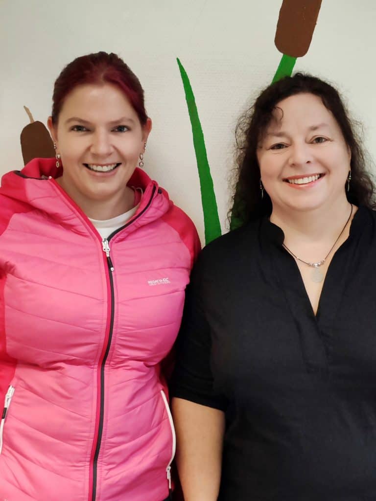 Zwei Frauen stehen nebeneinander und lächeln in die Kamera; die eine trägt eine leuchtend rosa Jacke und hat rote Haare, die andere ein schwarzes Oberteil und lockiges dunkles Haar. An der Wand hinter ihnen ist grüner Pflanzenschmuck zu sehen.