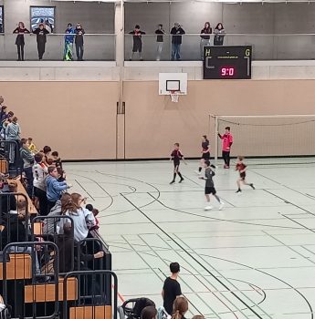 Kinder spielen ein Hallenfußballspiel in einer Turnhalle, während die Zuschauer von Tribünen und einem Balkon im Obergeschoss zusehen. Eine digitale Anzeigetafel zeigt den Spielstand an, und die Turnhalle ist mit Basketballkörben und Sportmarkierungen auf dem Boden ausgestattet.