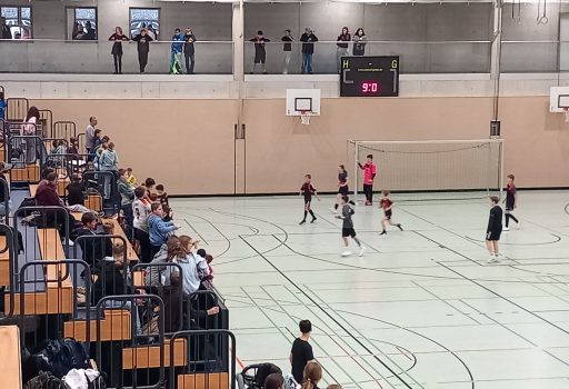 Kinder spielen ein Hallenfußballspiel in einer Turnhalle, während die Zuschauer von Tribünen und einem Balkon im Obergeschoss zusehen. Eine digitale Anzeigetafel zeigt den Spielstand an, und die Turnhalle ist mit Basketballkörben und Sportmarkierungen auf dem Boden ausgestattet.