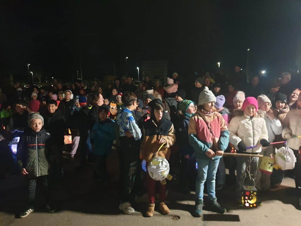 Eine große Gruppe von Kindern und Erwachsenen, viele in Wintermänteln und Mützen, versammelt sich nachts im Freien. Einige Kinder halten beleuchtete und geschmückte Laternen in der Hand, während sie zusammenstehen, was auf eine festliche oder gemeinschaftliche Veranstaltung hindeutet.