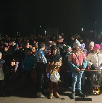 Eine große Gruppe von Kindern und Erwachsenen, viele in Wintermänteln und Mützen, versammelt sich nachts im Freien. Einige Kinder halten beleuchtete und geschmückte Laternen in der Hand, während sie zusammenstehen, was auf eine festliche oder gemeinschaftliche Veranstaltung hindeutet.