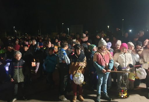 Eine große Gruppe von Kindern und Erwachsenen, viele in Wintermänteln und Mützen, versammelt sich nachts im Freien. Einige Kinder halten beleuchtete und geschmückte Laternen in der Hand, während sie zusammenstehen, was auf eine festliche oder gemeinschaftliche Veranstaltung hindeutet.