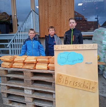 Drei Jungen stehen hinter einem Tisch, auf dem Brennholz vor einem Gebäude gestapelt ist. Auf einem Schild davor steht Schülerfirma, 5€ pro Sack, Bibrisschule, was bedeutet, dass sie Brennholz für 5 Euro pro Sack verkaufen.