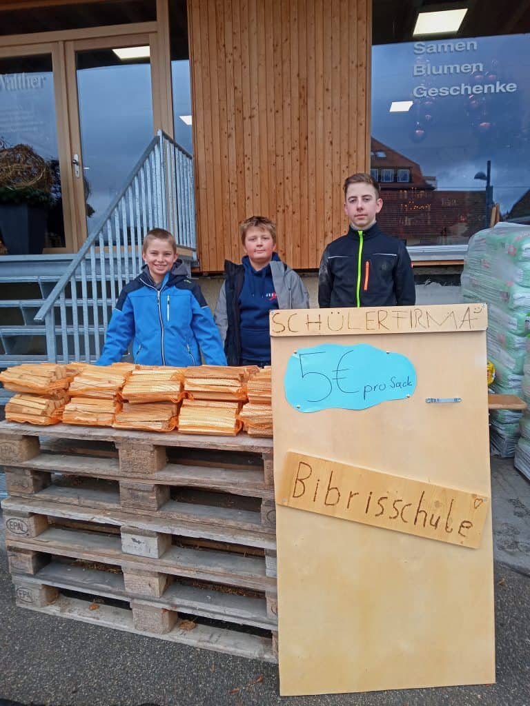 Drei Jungen stehen hinter einem Tisch, auf dem Brennholz vor einem Gebäude gestapelt ist. Auf einem Schild davor steht Schülerfirma, 5€ pro Sack, Bibrisschule, was bedeutet, dass sie Brennholz für 5 Euro pro Sack verkaufen.