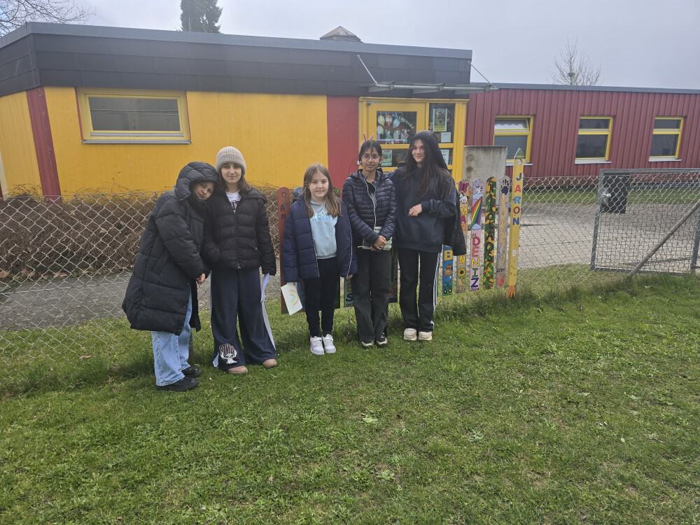 Fünf Mädchen stehen zusammen im Gras vor einem gelb-roten Gebäude. Sie tragen Jacken und Freizeitkleidung. In ihrer Nähe steht ein buntes Schild mit positiven Worten, neben einem Maschendrahtzaun.