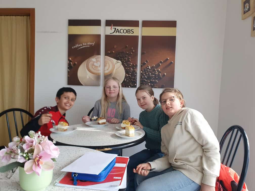 Vier Kinder sitzen um einen runden Tisch in einem gemütlichen Raum, lächeln und essen Kuchen. Hinter ihnen ist eine Wand mit drei Kunstwerken zum Thema Kaffee und einem Jacobs-Kaffee-Logo zu sehen. Auf dem Tisch steht ein rosa Blumengesteck.