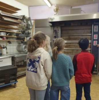 Vier Kinder stehen in einer Bäckereiküche und blicken auf einen großen Industrieofen. Auf den Regalen an der linken Wand sind Backgeräte und Backbleche zu sehen. Die Kinder stehen mit dem Rücken zur Kamera.