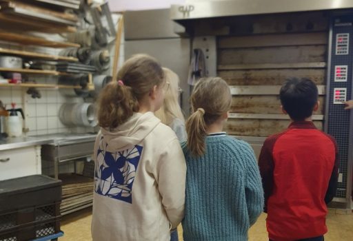 Vier Kinder stehen in einer Bäckereiküche und blicken auf einen großen Industrieofen. Auf den Regalen an der linken Wand sind Backgeräte und Backbleche zu sehen. Die Kinder stehen mit dem Rücken zur Kamera.