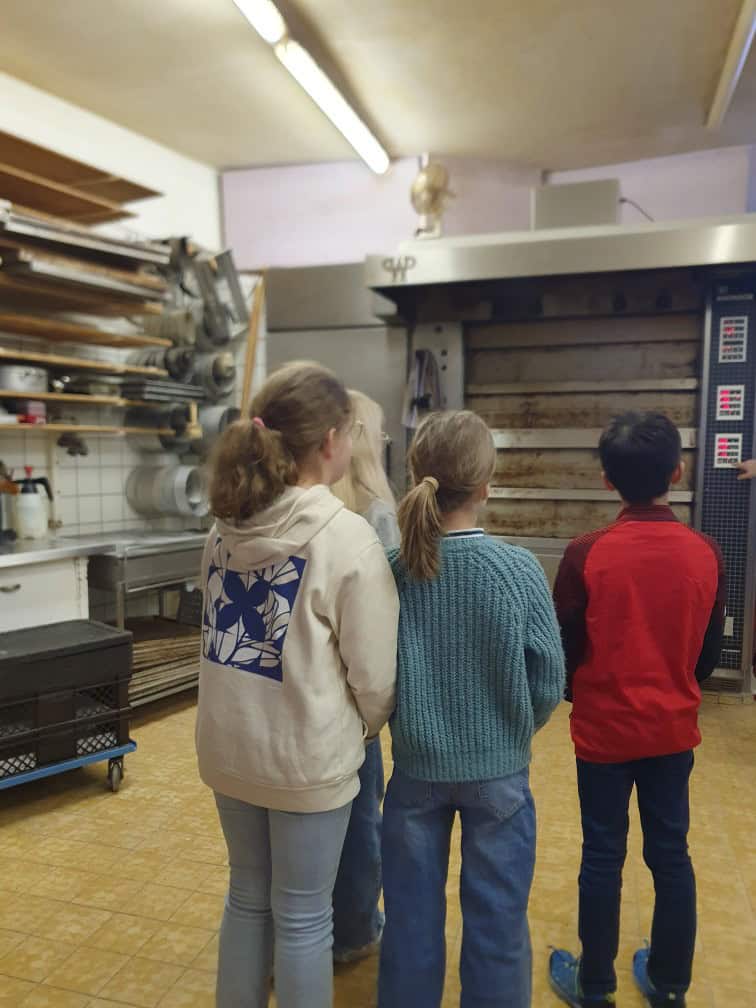 Vier Kinder stehen in einer Bäckereiküche und blicken auf einen großen Industrieofen. Auf den Regalen an der linken Wand sind Backgeräte und Backbleche zu sehen. Die Kinder stehen mit dem Rücken zur Kamera.