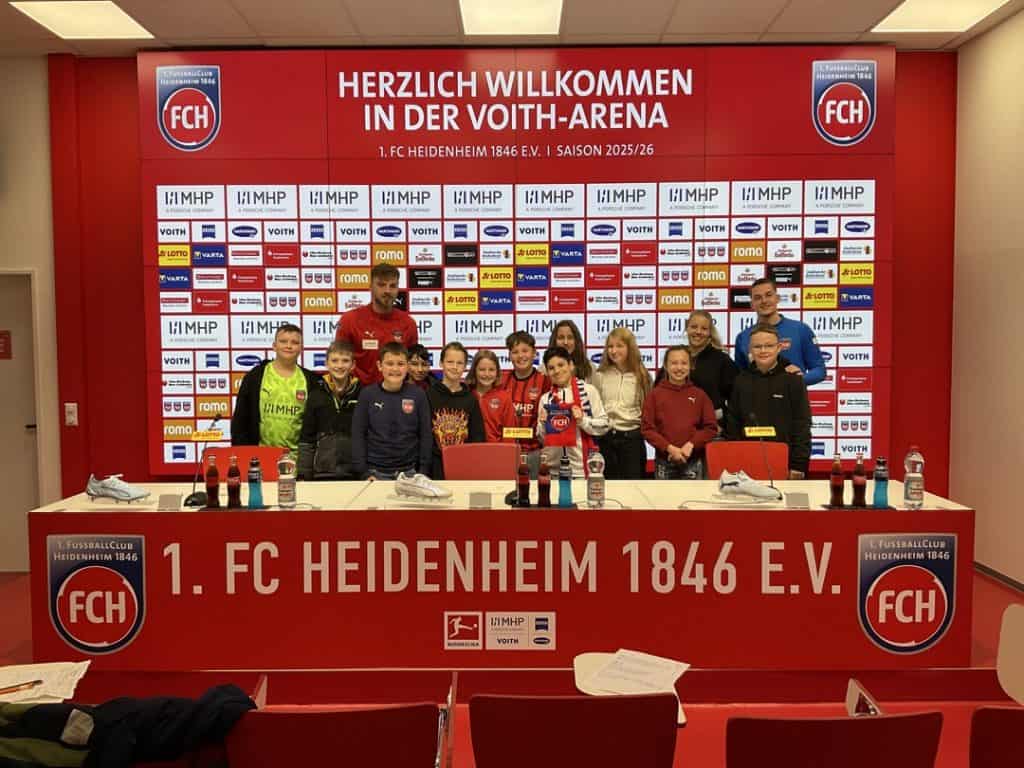 Eine Gruppe von Kindern und zwei Erwachsenen posieren vor einer Pressekulisse in der Voith-Arena des 1. FC Heidenheim 1846 e.V.. Sie lächeln, vor ihnen stehen Mikrofone und Getränke auf dem Tisch. Die Kulisse ist mit Sponsorenlogos bedeckt.