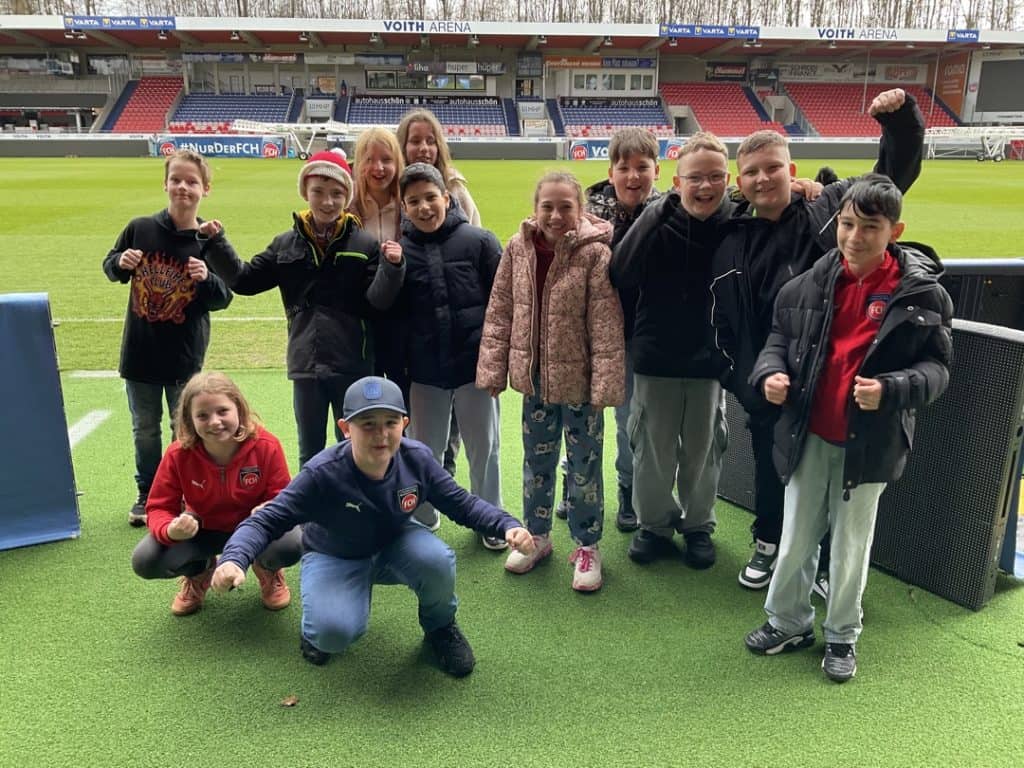 Eine Gruppe von zwölf Kindern in legerer Winterkleidung lächelt und posiert energisch am Rande eines Fußballstadions mit leeren roten und weißen Sitzen im Hintergrund.