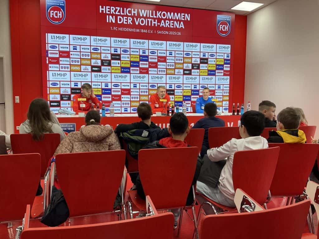 Eine Gruppe von Kindern sitzt vor einem Pressekonferenztisch, an dem drei Personen in roten FCH-Sporttrikots sitzen. Im Hintergrund sind die Logos der Sponsoren und ein Schild zu sehen, das die Besucher in der Voith-Arena begrüßt.