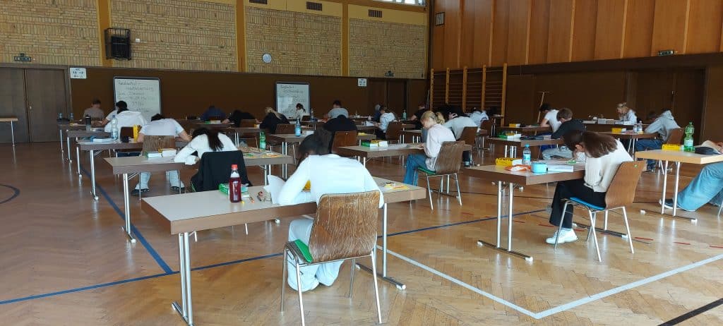 In einer geräumigen Turnhalle sitzen die Schüler an einzelnen Tischen und schreiben eine Prüfung. Sie sitzen mit Abstand zueinander, konzentriert auf ihre Unterlagen. Der Raum hat Holzwände und im Hintergrund sind Turngeräte zu sehen.