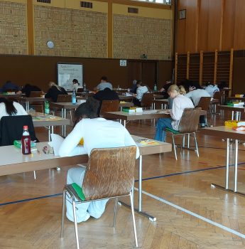 In einer geräumigen Turnhalle sitzen die Schüler an einzelnen Tischen und schreiben eine Prüfung. Sie sitzen mit Abstand zueinander, konzentriert auf ihre Unterlagen. Der Raum hat Holzwände und im Hintergrund sind Turngeräte zu sehen.
