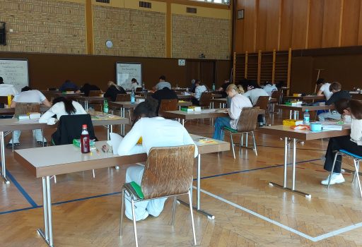 In einer geräumigen Turnhalle sitzen die Schüler an einzelnen Tischen und schreiben eine Prüfung. Sie sitzen mit Abstand zueinander, konzentriert auf ihre Unterlagen. Der Raum hat Holzwände und im Hintergrund sind Turngeräte zu sehen.