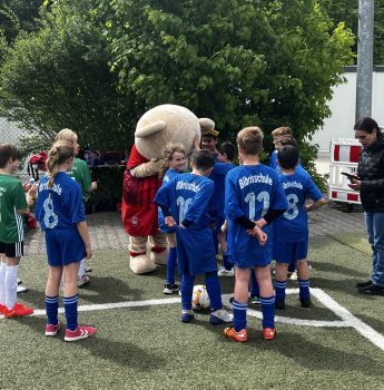Eine Gruppe von Kindern in blauen und grünen Fußballtrikots versammelt sich um eine Person in einem Bärenmaskottchenkostüm auf einem Fußballfeld, während ein Erwachsener in der Nähe steht. Im Hintergrund sind Bäume und ein Zaun zu sehen.