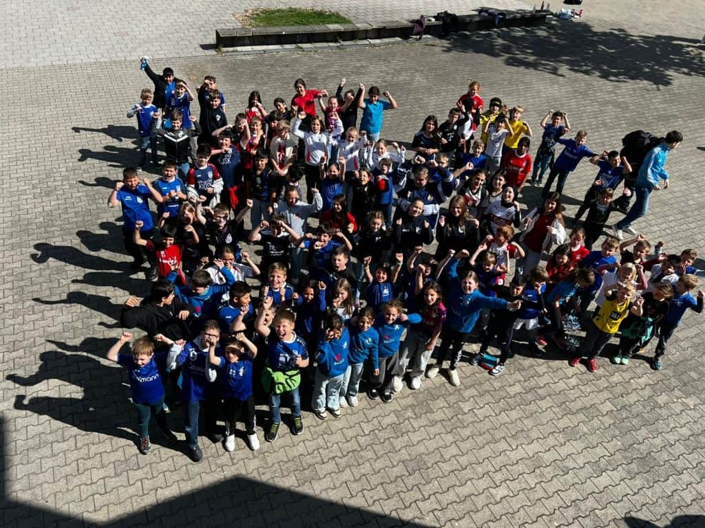 Eine große Gruppe von Kindern und einige Erwachsene stehen draußen auf einer gepflasterten Fläche zusammen, lächeln und winken in die Kamera, einige tragen bunte T-Shirts und heben aufgeregt die Arme.