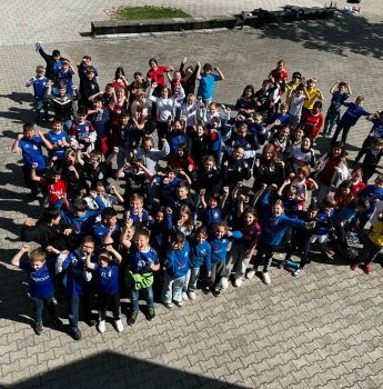 Eine große Gruppe von Kindern und einige Erwachsene stehen draußen auf einer gepflasterten Fläche zusammen, lächeln und winken in die Kamera, einige tragen bunte T-Shirts und heben aufgeregt die Arme.