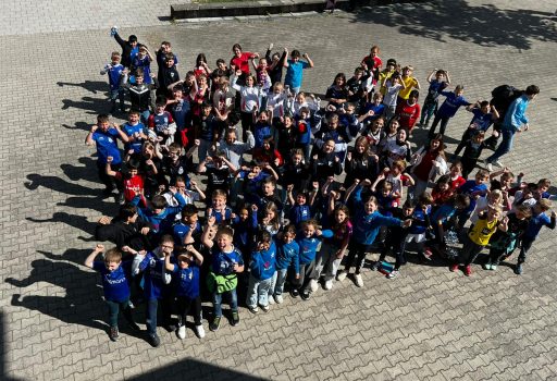 Eine große Gruppe von Kindern und einige Erwachsene stehen draußen auf einer gepflasterten Fläche zusammen, lächeln und winken in die Kamera, einige tragen bunte T-Shirts und heben aufgeregt die Arme.