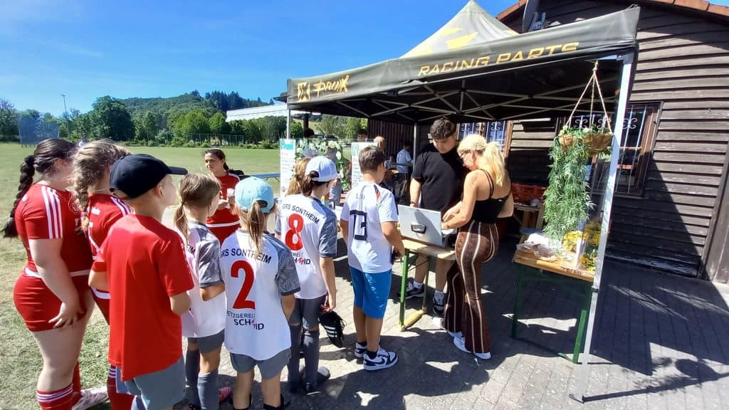 Eine Gruppe von Kindern in Sportuniformen steht an einem sonnigen Tag vor einem Zelt mit der Aufschrift „Racing Parts“ in einer Reihe, wo zwei Erwachsene an einem Tisch mit einem Laptop und verschiedenen Gegenständen arbeiten, direkt neben einem Holzgebäude.