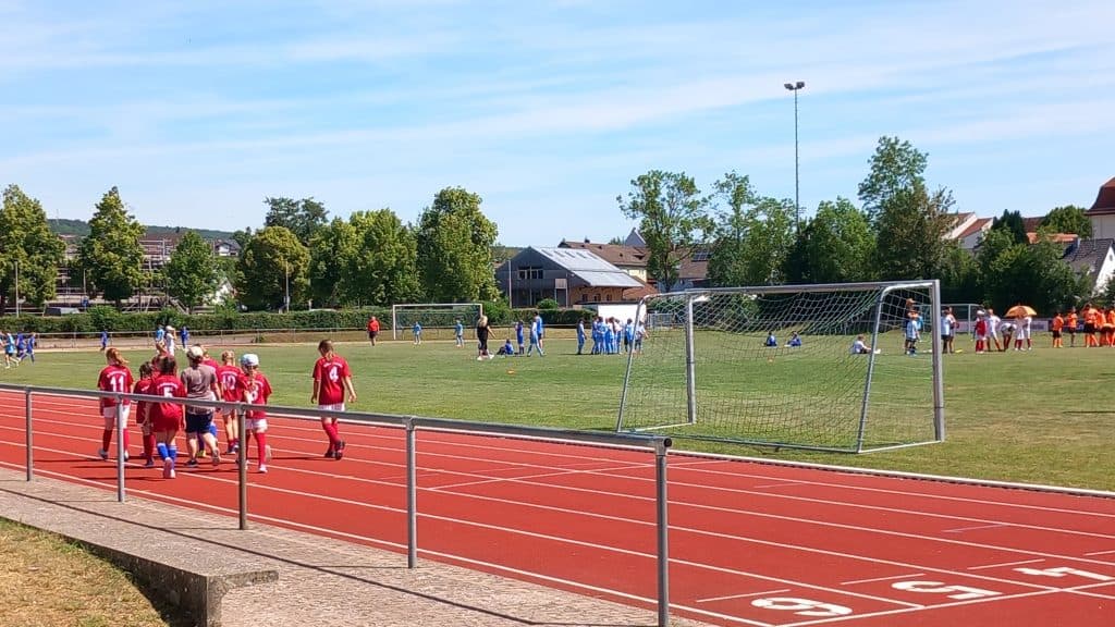 Kinder in roten Fußballtrikots laufen auf einer Laufbahn in der Nähe eines Fußballplatzes entlang, auf dem Kinder in blauen und weißen Trikots spielen, während auf der anderen Seite eine weitere Gruppe in orangefarbenen Trikots zu sehen ist. Im Hintergrund sind Bäume und Häuser zu erkennen.