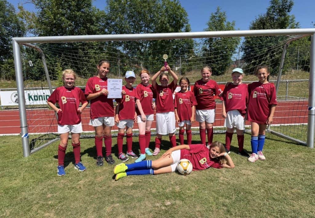 Neun Mädchen in roten Fußballtrikots posieren vor einem Tor und halten einen Pokal und eine Urkunde in den Händen. Ein Mädchen liegt mit einem Fußball vor dem Tor auf dem Rasen. Die Szene spielt sich bei sonnigem Wetter ab, im Hintergrund sind Bäume zu sehen.