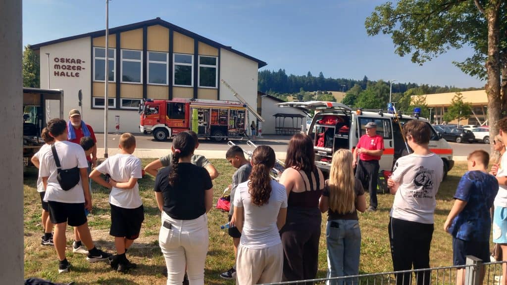 An einem sonnigen Tag hört eine Gruppe von Kindern und Jugendlichen den Feuerwehrleuten zu, die vor einem Gebäude mit der Aufschrift „Oskar-Mozer-Halle“ in der Nähe der Löschfahrzeuge ihre Ausrüstung vorführen.