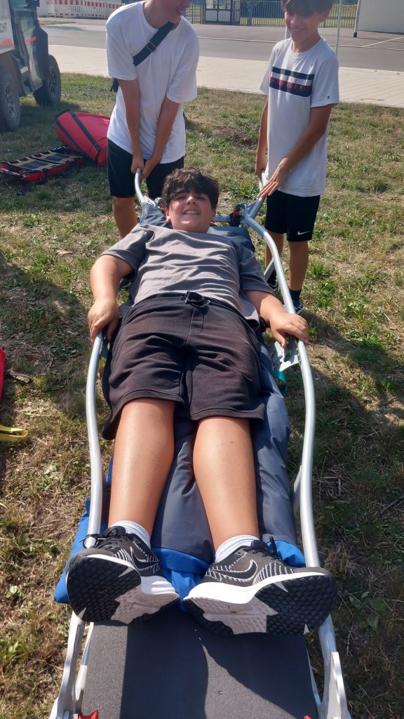 Ein Junge liegt lächelnd auf einer Trage im Freien, während zwei andere Jungen in weißen Hemden und Shorts die Griffe der Trage festhalten und sich darauf vorbereiten, ihn an einem sonnigen Tag anzuheben oder über eine Rasenfläche zu tragen.