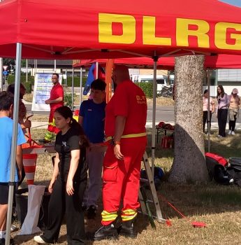 Bei einer Veranstaltung im Freien steht eine Gruppe von Menschen unter und um ein rotes Zelt mit der Aufschrift „DLRG“. Einige tragen rote Uniformen, andere Freizeitkleidung. Unter dem Zelt sind Ausrüstung und Schilder zu sehen, und im Hintergrund sind weitere Personen zu erkennen.