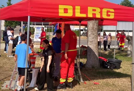 Bei einer Veranstaltung im Freien steht eine Gruppe von Menschen unter und um ein rotes Zelt mit der Aufschrift „DLRG“. Einige tragen rote Uniformen, andere Freizeitkleidung. Unter dem Zelt sind Ausrüstung und Schilder zu sehen, und im Hintergrund sind weitere Personen zu erkennen.