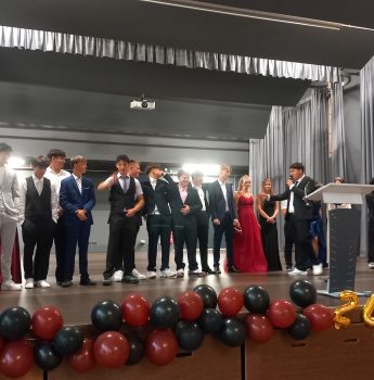 Eine Gruppe junger Menschen in festlicher Kleidung steht auf einer mit roten und schwarzen Luftballons sowie einem Schild mit der Aufschrift „2024“ geschmückten Bühne und nimmt an einer Veranstaltung teil, die offenbar ein Abschlussball oder ein Abschlussfest ist.