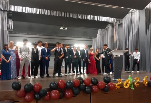 Eine Gruppe junger Menschen in festlicher Kleidung steht auf einer mit roten und schwarzen Luftballons sowie einem Schild mit der Aufschrift „2024“ geschmückten Bühne und nimmt an einer Veranstaltung teil, die offenbar ein Abschlussball oder ein Abschlussfest ist.