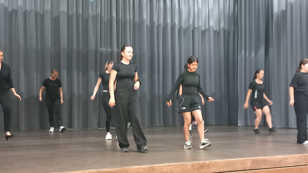 Eine Gruppe von Menschen in schwarzer Kleidung probt auf einer Bühne vor grauen Vorhängen eine Tanzchoreografie. Zwei Personen stehen vorne und führen die Gruppe an.