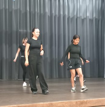 Eine Gruppe von Menschen in schwarzer Kleidung probt auf einer Bühne vor grauen Vorhängen eine Tanzchoreografie. Zwei Personen stehen vorne und führen die Gruppe an.