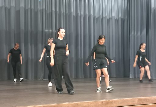 Eine Gruppe von Menschen in schwarzer Kleidung probt auf einer Bühne vor grauen Vorhängen eine Tanzchoreografie. Zwei Personen stehen vorne und führen die Gruppe an.