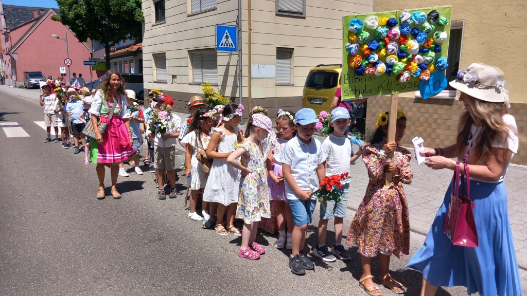 Eine Gruppe von Kindern in bunten Hüten und Sommerkleidung steht in einer Reihe auf einer sonnigen Straße, angeführt von Frauen in Kleidern. Ein Kind hält ein Schild in der Hand, das mit Papierblumen verziert ist. Im Hintergrund sind Gebäude und geparkte Autos zu sehen.