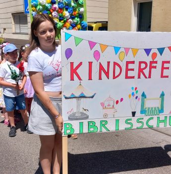 Eine Gruppe von Kindern steht draußen in einer Reihe und hält ein buntes Schild mit der Aufschrift „Kinderfest Bibrisschule“ hoch, das mit Zeichnungen von Luftballons, einem Riesenrad und Karnevalsszenen verziert ist. Die Sonne scheint strahlend.